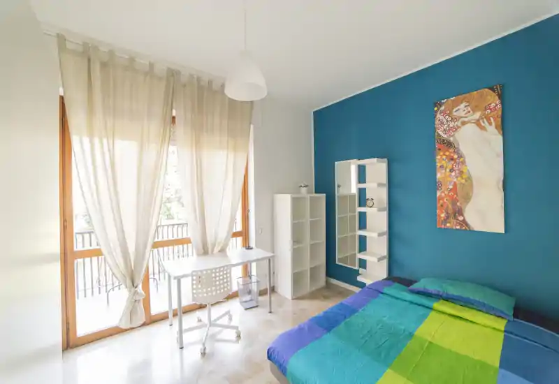 Bright, modern bedroom with a double bed, colorful bedding, a desk by a large balcony window and shelving against a teal accent wall.