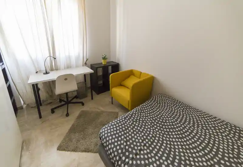 Bright, compact bedroom with a patterned single bed, a white desk by the window and a cheerful yellow armchair. Natural light fills the room, which includes a small rug and minimal shelving — ideal for a student or single occupant.