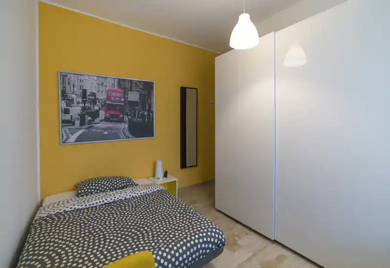 Compact, well-styled bedroom with a polka-dot duvet, warm yellow accent wall and large white wardrobe. The space is bright and cozy, ideal for a single occupant or short stays.