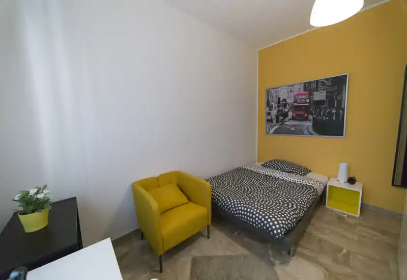 Bright, cozy bedroom with a double bed, patterned bedding and a mustard-yellow accent wall. A matching yellow armchair and bedside table add style and practical seating.