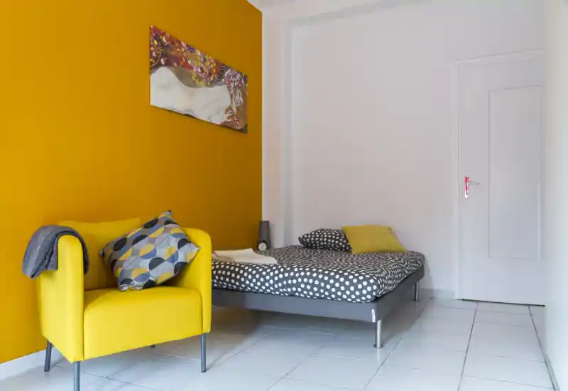 Camera da letto luminosa e moderna con letto a pois e una poltrona gialla vivace davanti a una parete d'accento ocra. Linee pulite e cuscini coordinati rendono lo spazio accogliente e perfetto per un annuncio per soggiorni brevi.