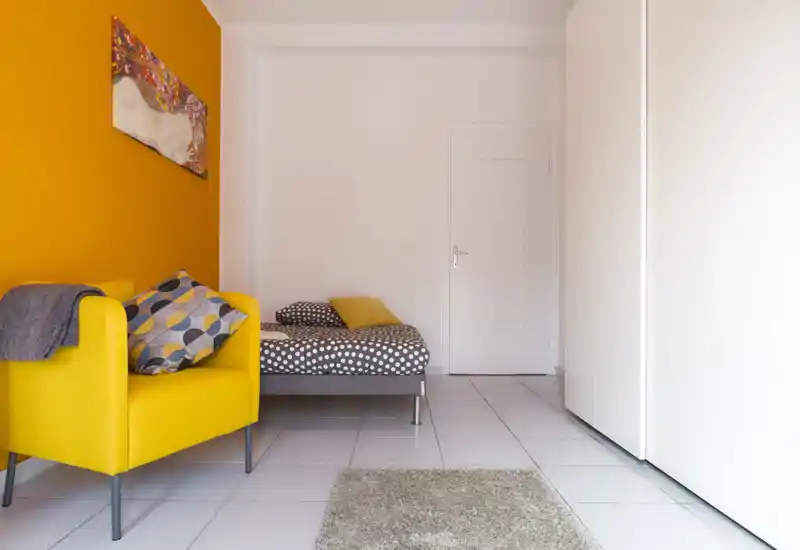 Camera da letto compatta e stilosa con letto a pois, poltrona gialla vivace e parete d'accento arancione che crea un'atmosfera accogliente e moderna.