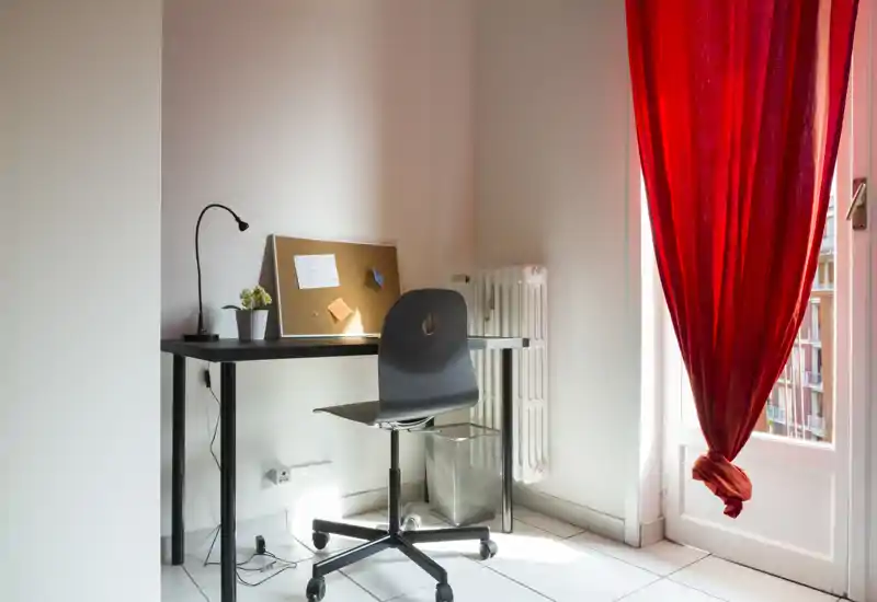 Angolo camera usato come area lavoro con scrivania essenziale, sedia, bacheca e luce naturale che entra dalla porta con tenda rossa.