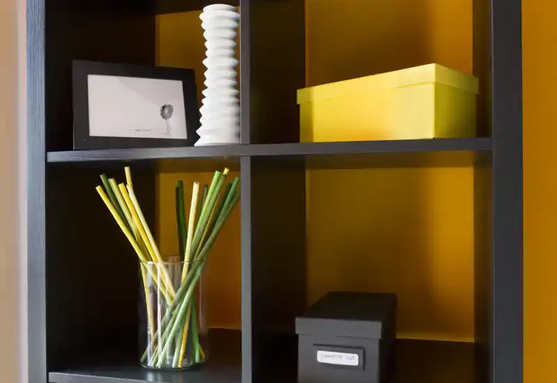 Primo piano di una scaffalatura nera a cubi con oggetti decorativi: un vaso di vetro con bastoncini verdi e gialli, una stampa incorniciata, un vaso scultoreo bianco e due scatole portaoggetti su sfondo giallo-arancione.