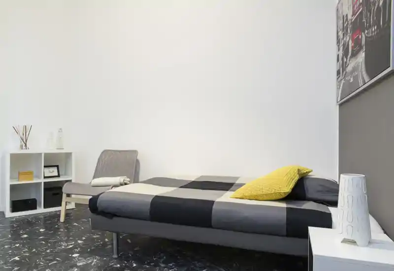 Dormitorio moderno y minimalista con cama baja, edredón gris de cuadros y cojín amarillo; paredes blancas y estantería sencilla que crean una atmósfera contemporánea y luminosa.