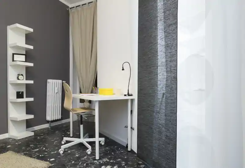 Rincón compacto de estar/trabajo con un pequeño escritorio blanco, silla con ruedas y estantería minimalista. Tonos neutros y cortinas suaves crean un espacio limpio y funcional, ideal para estudiar o trabajar desde casa.