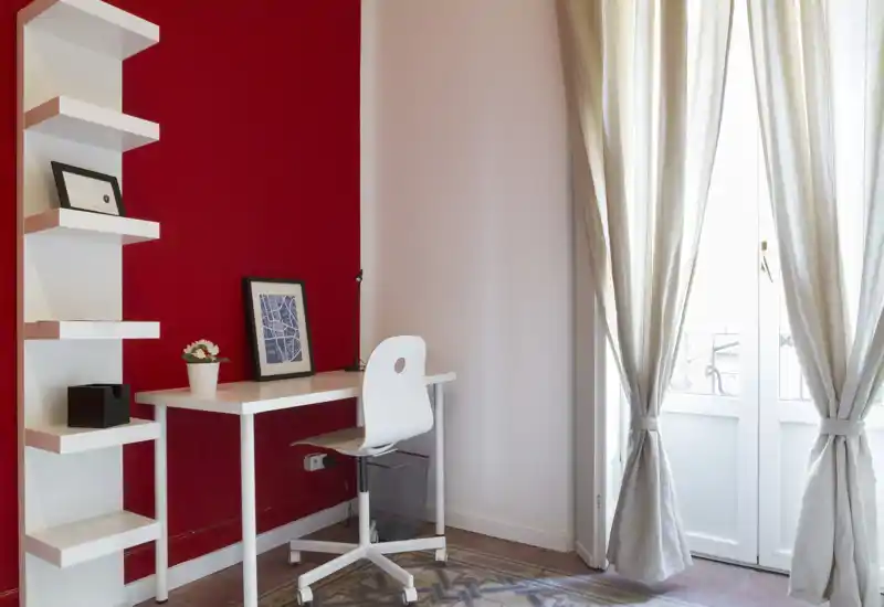 Angolo studio luminoso e ordinato con scrivania e sedia bianche davanti a una parete rossa d'accento e tende chiare che lasciano entrare luce naturale.