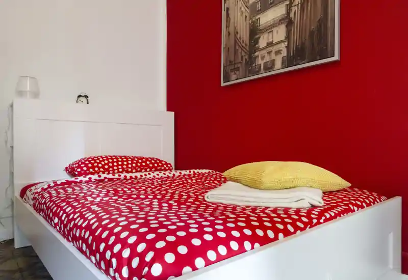 Camera da letto luminosa e moderna con letto bianco e parete rossa a contrasto. La trapunta a pois e i cuscini stratificati creano un’atmosfera accogliente e contemporanea.