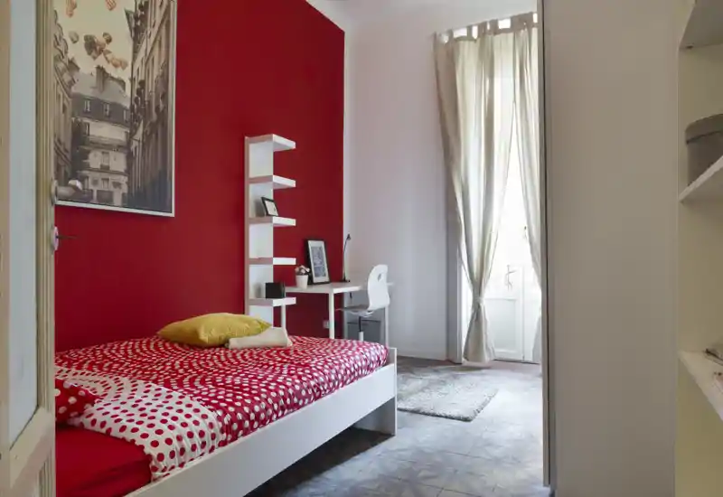 Camera da letto luminosa e stilosa con letto bianco e copriletto rosso a pois, una scrivania compatta e una finestra alta che lascia entrare luce naturale—accogliente e ben arredata.