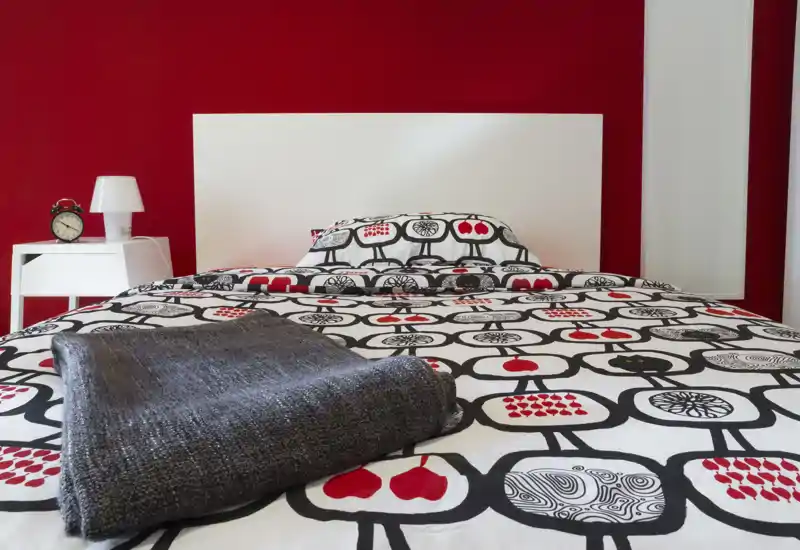 Dormitorio luminoso y bien ambientado con colcha estampada, cabecero blanco y manta gris doblada. La pared roja y la mesita ordenada crean un ambiente moderno y acogedor, perfecto para un anuncio de alquiler.