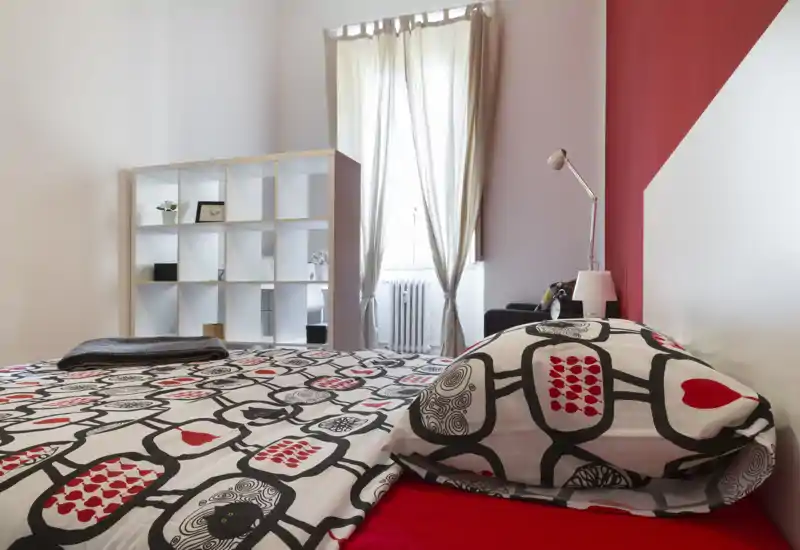 Dormitorio luminoso y con estilo, con ropa de cama estampada, una lámpara de mesilla y luz natural de una ventana alta; estanterías ordenadas aportan almacenamiento e interés visual.