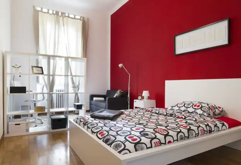 Dormitorio luminoso y moderno con cama blanca, ropa de cama estampada y una pared roja llamativa; la luz natural inunda la estancia y una estantería divide la zona de trabajo y el sofá.
