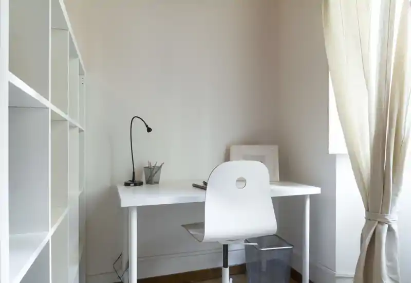 Rincón de dormitorio luminoso y minimalista usado como pequeña zona de estudio con mesa blanca, silla, lámpara y estanterías junto a una ventana con cortina.
