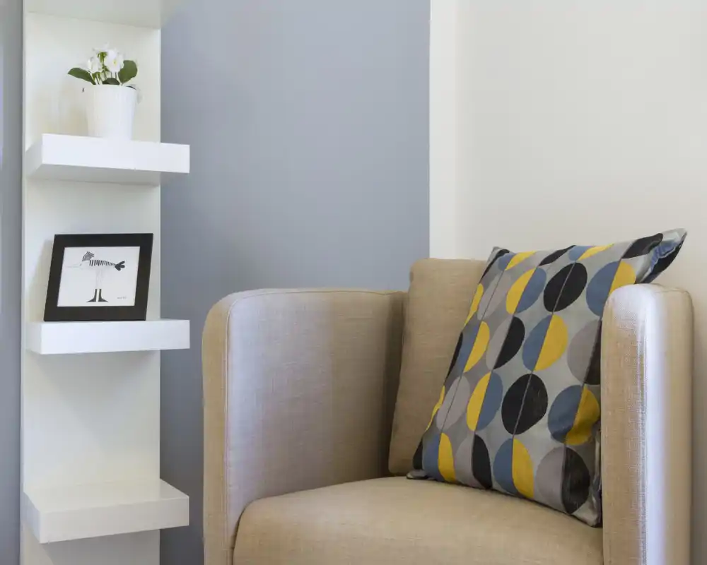 Angolo accogliente con poltrona beige imbottita, cuscino decorato e mensole minimal con oggetti: area giorno luminosa e curata, perfetta per un annuncio.