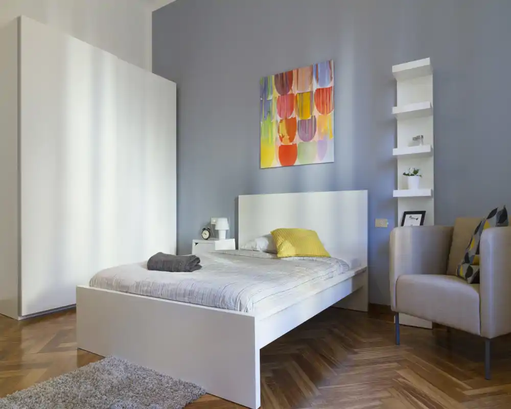 Camera da letto moderna e luminosa con letto bianco, quadri colorati e una poltrona accogliente — perfetta per un soggiorno in città.