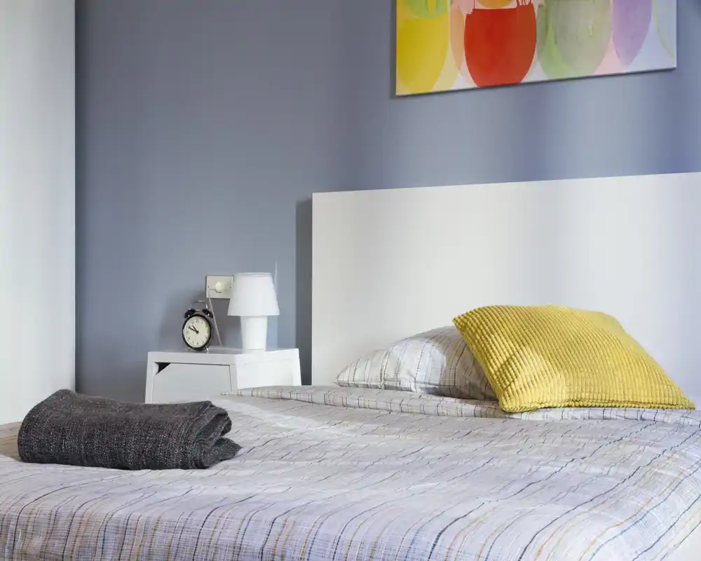 Camera da letto moderna e luminosa con letto matrimoniale ben rifatto, cuscino giallo e comodino con lampada e sveglia — arredo pulito e funzionale adatto per annunci.