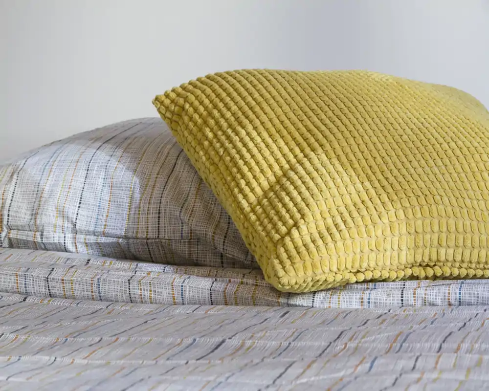 Primo piano di un letto ben rifatto con un cuscino giallo testurizzato e biancheria a fantasia; dettagli nitidi, luminosi e accoglienti ideali per un annuncio di camera da letto.