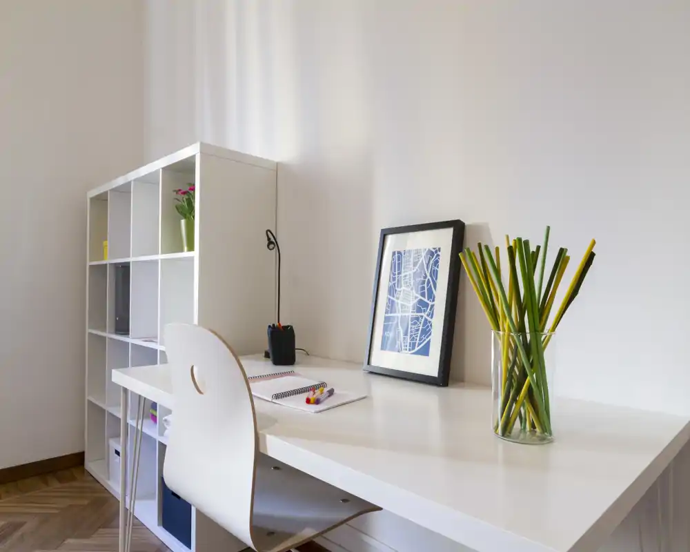 Angolo studio luminoso e minimal in soggiorno: scrivania bianca con sedia, quadro, lampada da tavolo e vaso decorativo con steli. Linee pulite e luce naturale rendono l'area accogliente.