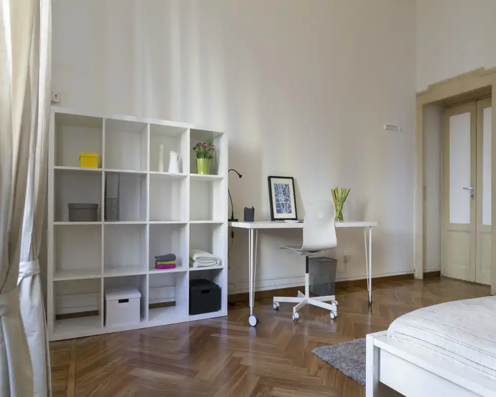 Angolo letto luminoso e minimal con mobile a cubi bianco, scrivania ordinata e parte del letto; parquet pulito e luce naturale donano un’atmosfera ariosa e moderna.