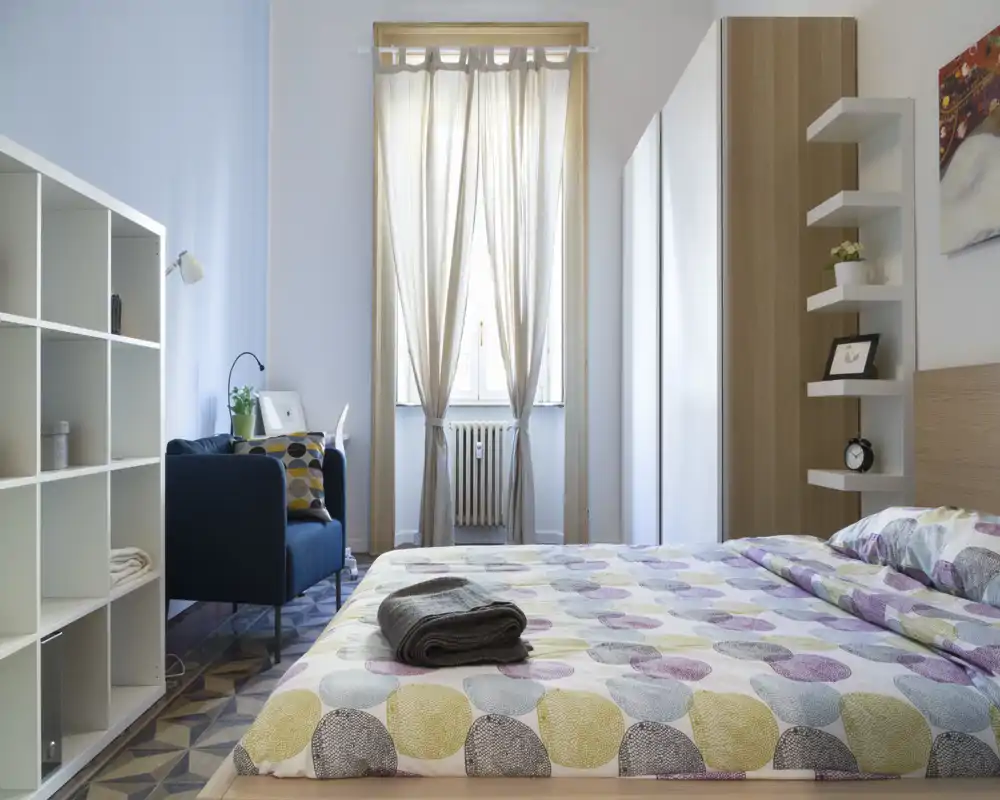 Bright, modern bedroom with a patterned duvet and a large bed in the foreground; a cozy seating area and shelving add functional charm.