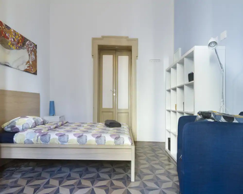 Bright, tidy bedroom with a wooden bed, patterned duvet and a minimalist shelving unit. The room features decorative artwork, a bedside lamp and geometric floor tiles.