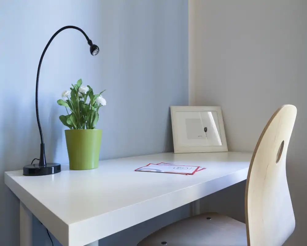Bright, minimal desk area with a white work surface, adjustable black desk lamp, a small green potted plant and a light wooden chair — ideal for a compact home office or study corner.