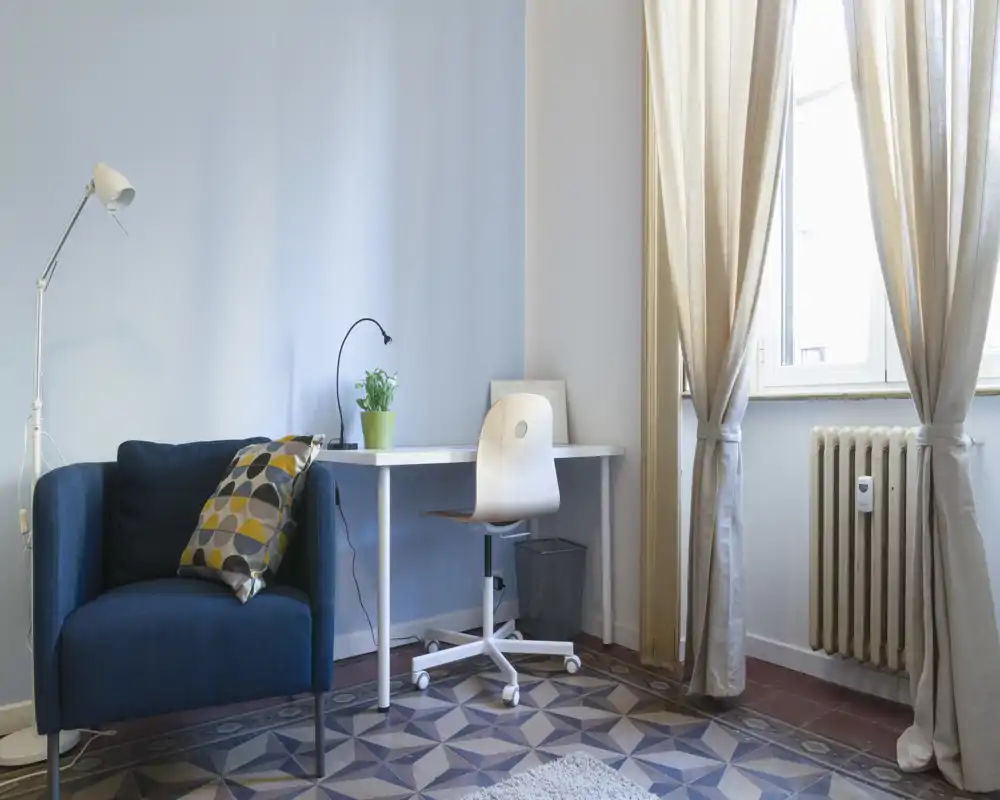 Bright, stylish living corner with a navy armchair, patterned cushion, desk and natural light from a large window — ideal for relaxing or working.