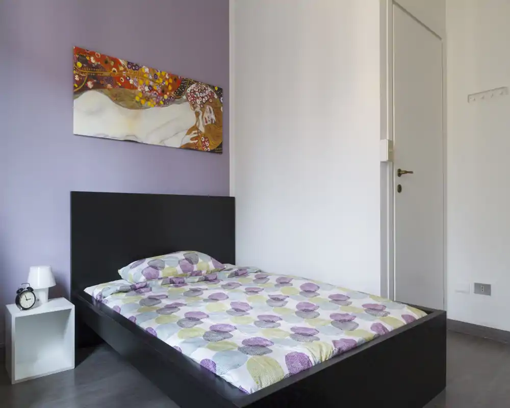 Camera da letto luminosa e minimale con letto matrimoniale, biancheria a fantasia, comodino con lampada e quadro a parete che creano un’atmosfera accogliente.