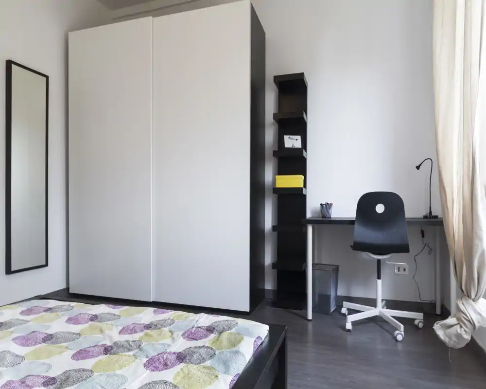 Camera da letto moderna con grande armadio, vista parziale del letto con copriletto a fantasia e una piccola scrivania vicino alla finestra. Luminosa, minimal e ordinata — ideale per singoli o studenti.