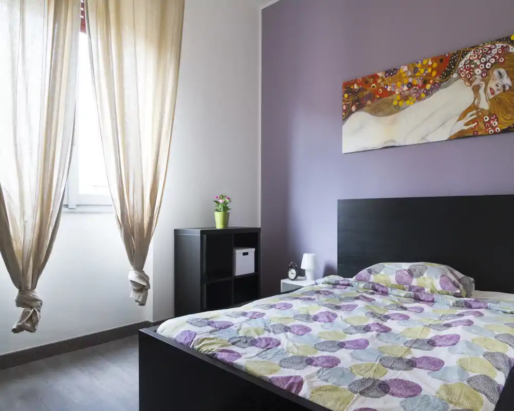 Camera da letto moderna e luminosa con letto matrimoniale, biancheria a fantasia, testata scura e luce naturale morbida da una finestra con tende leggere. Sulla parete è presente un quadro colorato; un mobiletto e un comodino offrono spazio contenitivo.