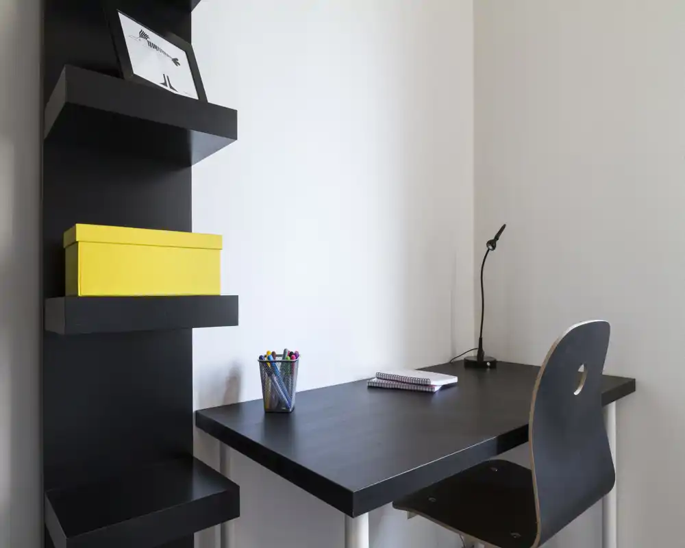 Piccolo angolo studio minimalista con mensole nere a parete, scrivania e sedia abbinate nere, lampada da tavolo e materiale di cancelleria — un angolo di lavoro moderno e ordinato per una persona.