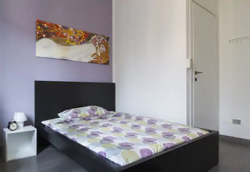 Dormitorio luminoso y sencillo con cama doble, ropa de cama estampada, mesita con lámpara y cuadro en la pared que crean un ambiente acogedor.