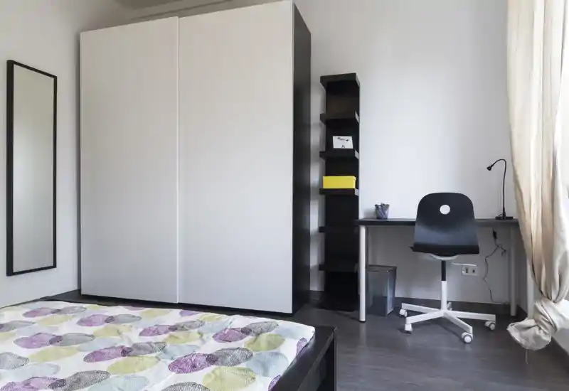 Dormitorio moderno con gran armario, vista parcial de la cama con edredón estampado y un pequeño espacio de trabajo junto a la ventana. Luminoso, minimalista y ordenado — ideal para una persona o estudiante.