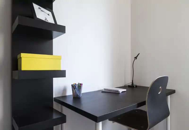 Pequeño espacio de trabajo minimalista con estanterías negras, escritorio y silla a juego, lámpara de mesa y material de papelería: un rincón de estudio moderno y ordenado para una persona.
