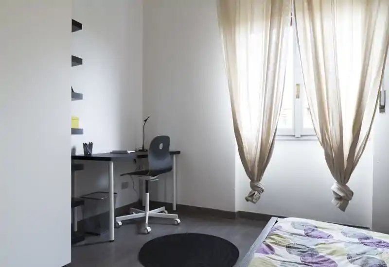 Rincón de dormitorio luminoso y minimalista con una cama individual, ropa de cama estampada y un pequeño escritorio junto a la ventana que ofrece un práctico espacio de estudio.