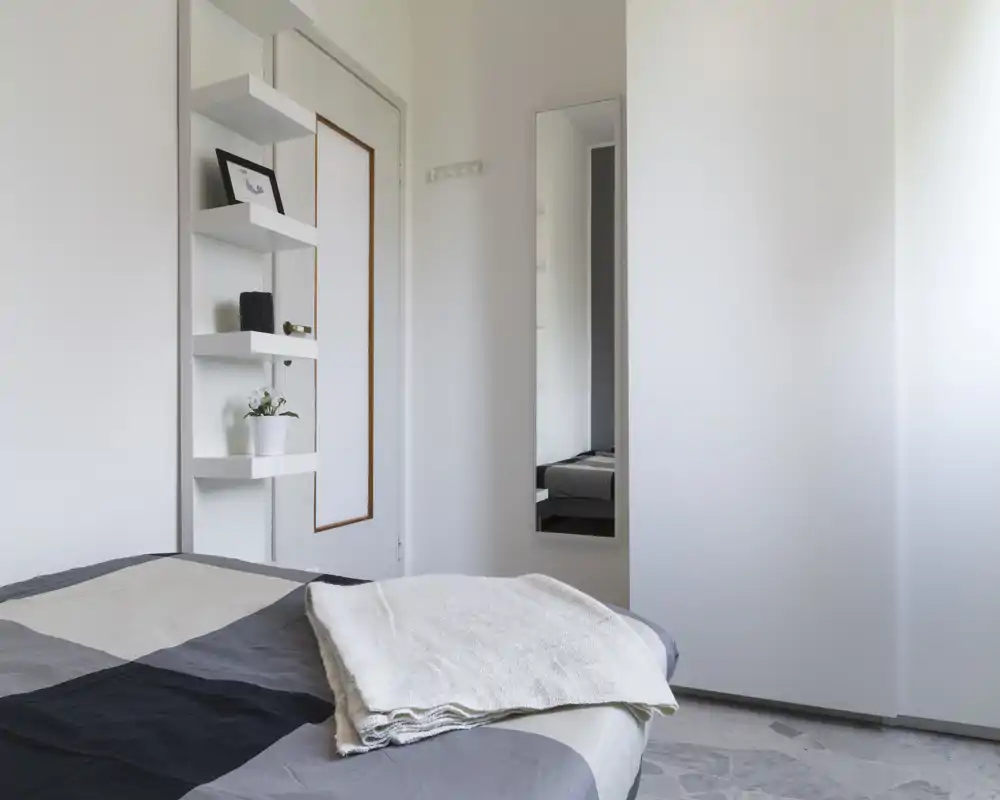 Minimalist bright bedroom with a neatly made double bed, soft linens and a wall mirror that reflects part of the room. Clean white walls, open shelving and natural light make this room feel calm and airy — ideal for rental photos.