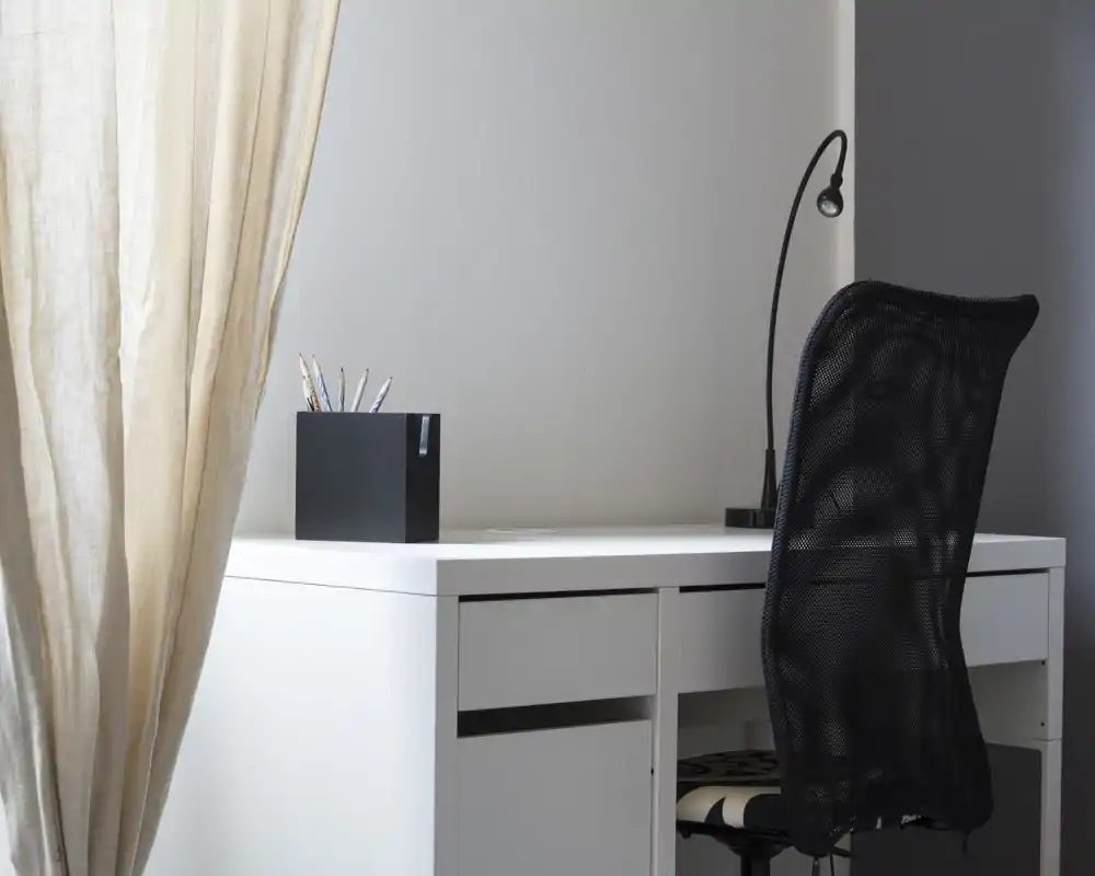 Bright, minimal home office corner with a white desk, mesh desk chair, desk lamp and pencil holder beside a soft curtain — a calm workspace with clean lines.