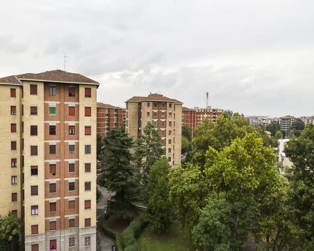 Exterior view showing residential apartment blocks and a tree-lined green area under an overcast sky; suitable as a terrace or balcony photo highlighting the neighborhood and greenery.