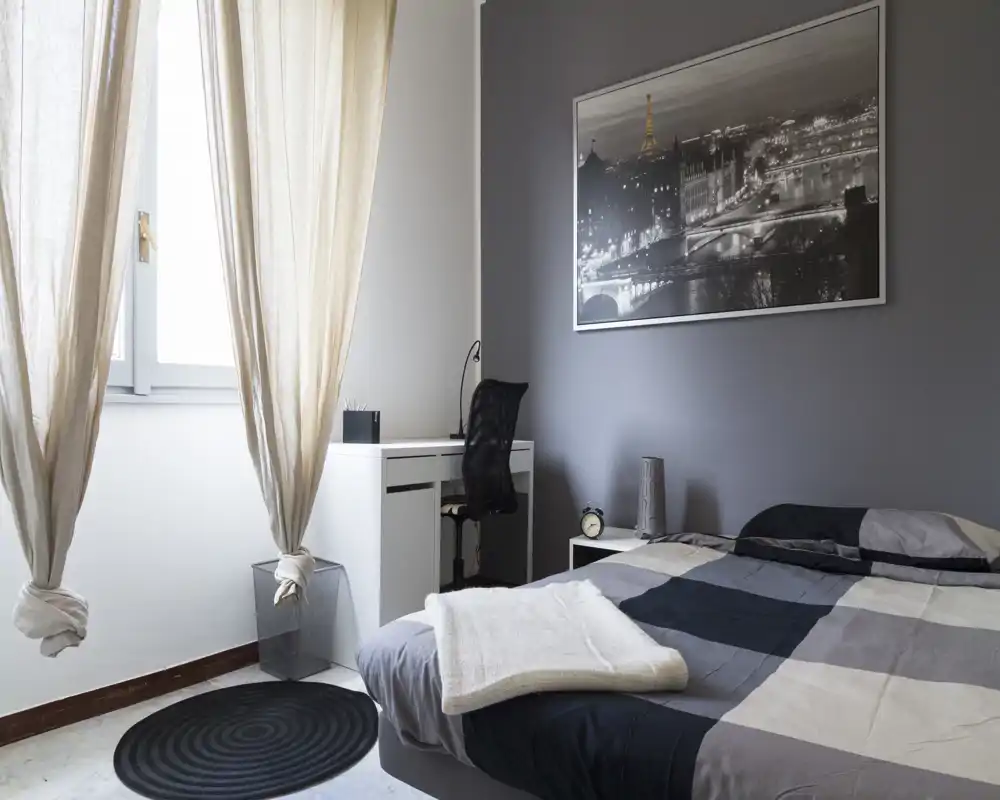 Bright, modern bedroom with a neatly made double bed, bedside table and a small desk by the window. Neutral gray tones and soft natural light create a calm, minimalist atmosphere ideal for rentals.