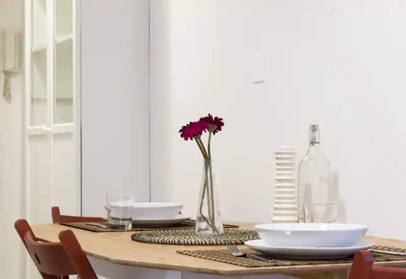 Área de comedor con mesa de madera para dos, salvamanteles de fibra y un jarrón delgado con flores. Luminoso, minimalista y acogedor para comidas informales.
