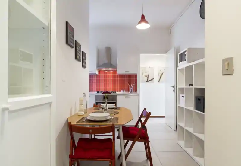 Cocina moderna y compacta con salpicadero de azulejos rojos, pequeña mesa de comedor y dos sillas acolchadas — luminosa, bien presentada y perfecta para una pareja o persona sola.