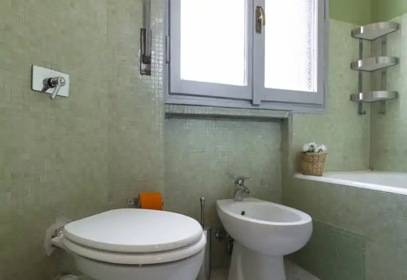 Baño compacto con inodoro, bidé y bañera blancos sobre azulejos de mosaico verde claro; ventana translúcida aporta luz suave.