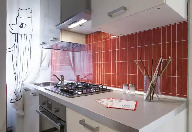 Cocina moderna y luminosa con placa de gas, fregadero de acero inoxidable y llamativa pared de azulejos rojos; encimera ordenada con utensilios y cortina decorativa.