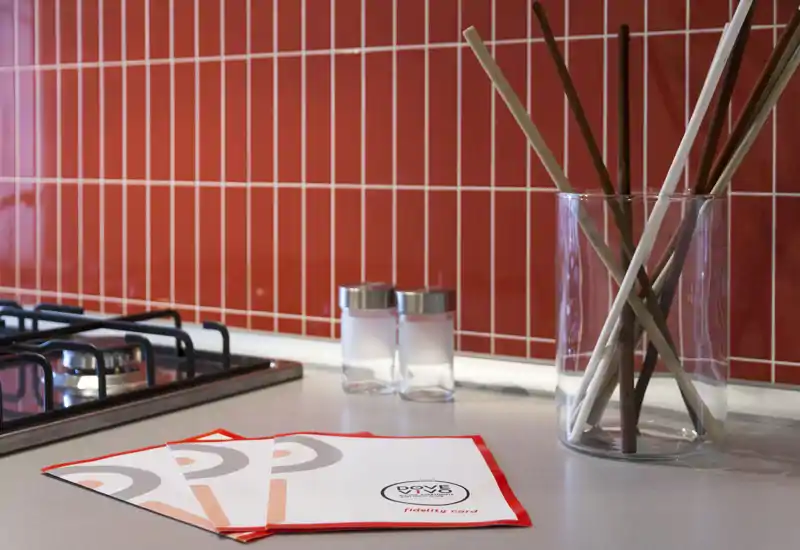 Plano detalle de una encimera de cocina moderna con placa de gas, salpicadero de azulejos rojos, jarrón decorativo de vidrio y pequeños salseros — una zona de preparación ordenada y estilizada.