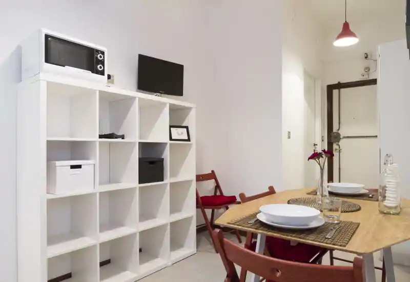 Rincón de comedor acogedor con mesa de madera puesta para dos, sillas plegables rojas, manteles individuales y un pequeño jarrón con flores — luminoso y de estilo sencillo para un apartamento compacto.