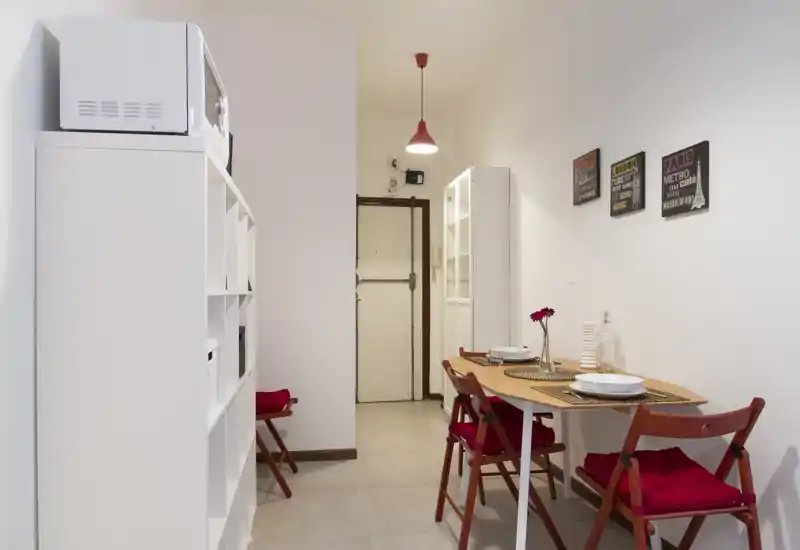 Zona de comedor compacta con mesa de madera puesta para dos y sillas plegables con cojines rojos; decoración sencilla y luminosa con cuadros en la pared y una lámpara colgante, acogedora para comer.