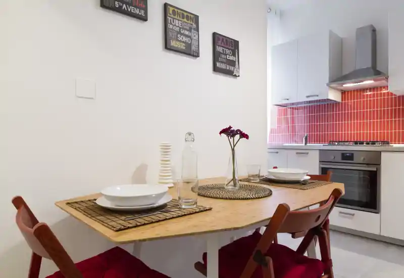Acogedor comedor en cocina abierta con mesa de madera para dos, cojines rojos y salpicadero de azulejos rojos. Luminoso, moderno y bien dispuesto — perfecto para anuncios de alquiler.