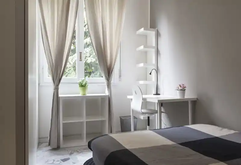 Camera da letto luminosa e minimalista con letto visibile in primo piano, piccolo scrittoio bianco e libreria vicino a una finestra illuminata con tende neutre e piante in vaso.