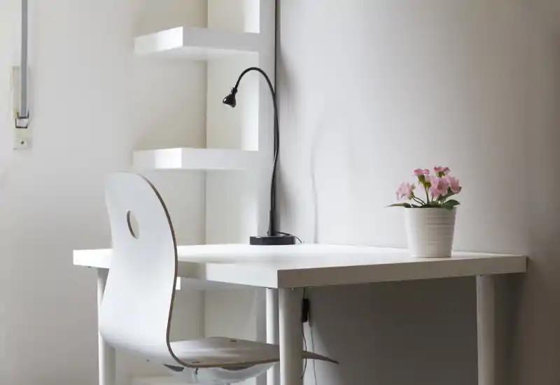 Angolo studio luminoso e minimal con una scrivania bianca, sedia moderna, lampada da scrivania e una piccola pianta in vaso — perfetto per un ufficio domestico compatto.