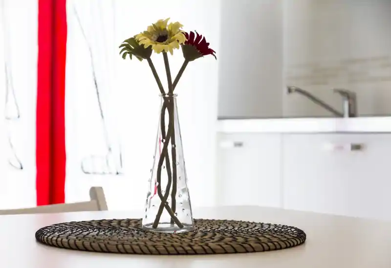 Surface de cuisine/salle à manger lumineuse avec un vase en verre et des fleurs sur un dessous de table tressé, prise avec un arrière-plan doux et lumière naturelle.